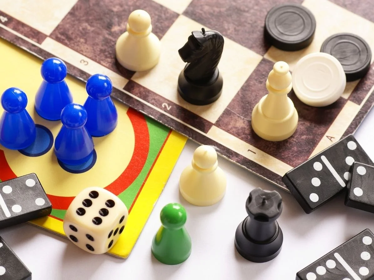 A game of chess and dice with pieces on a table. The chessboard has black and white pieces, and there are blue, green, black, and white game pieces around a second game board, likely for a different game. Several black and white dice are also present.