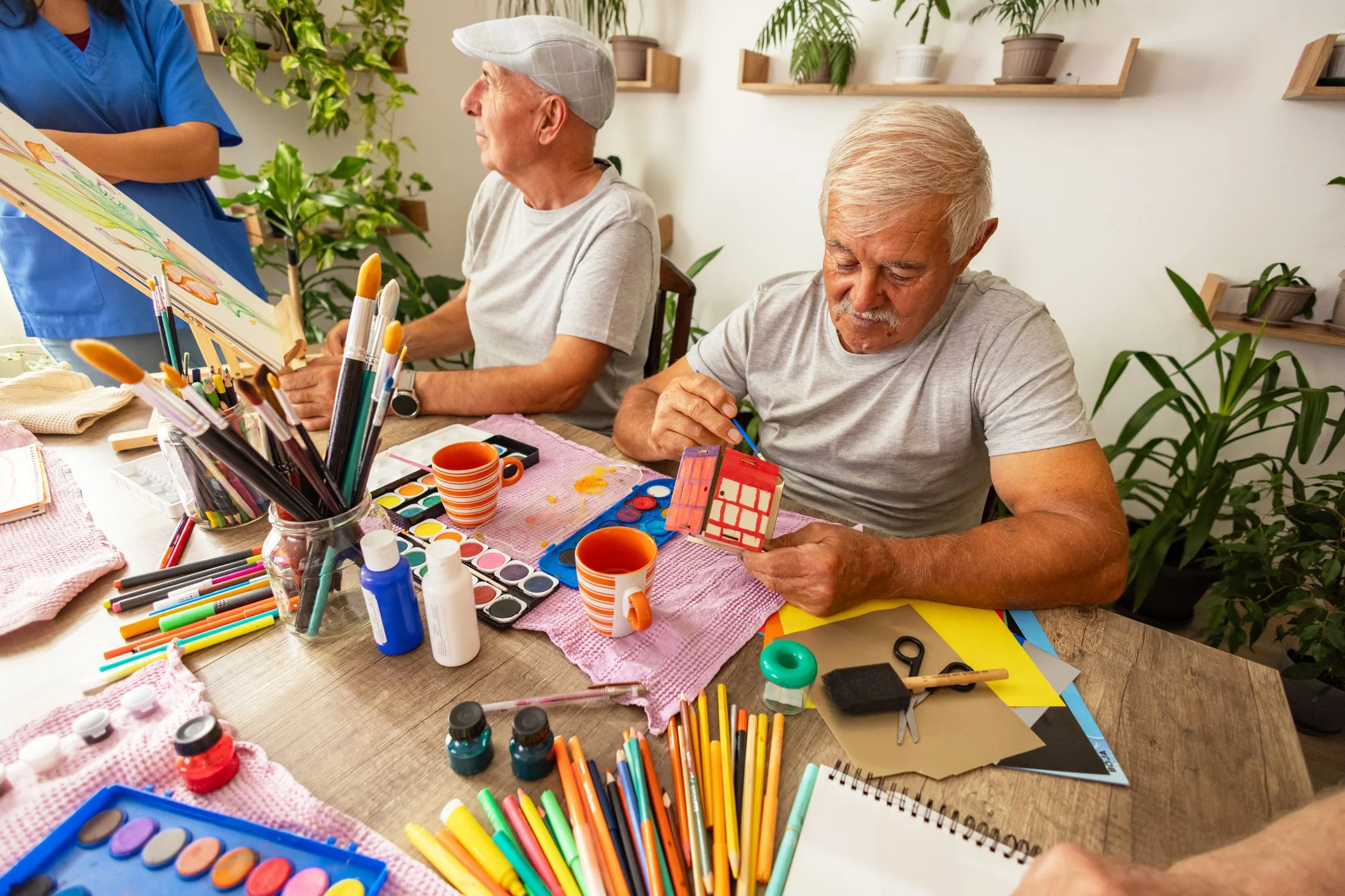 Two elderly men sit at a table engaged in arts and crafts activities. One man is holding a small, painted object resembling a miniature house, while the other is looking at a colorful artwork. The table is scattered with paints, markers, brushes, and craft supplies in front of them, with cups and bottles of paint nearby. A woman stands beside them, partially visible, assisting or observing. The room is decorated with green plants on the walls and shelves.