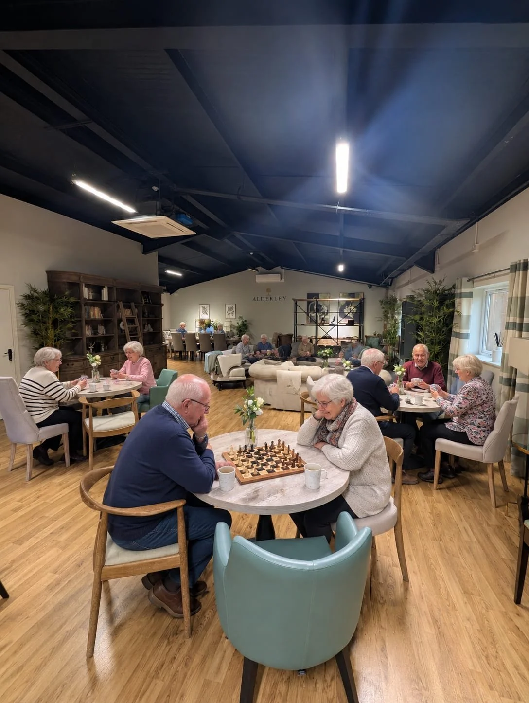 A group of elderly people playing chess at a round table in a cozy, well-lit communal lounge with wooden floors, bookshelves, and plants.