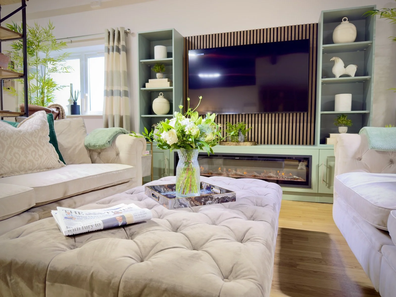 Living room with beige tufted sofa, glass vase with white flowers on a tufted ottoman, TV on a wall with shelves, electric fireplace below TV, window with striped curtains, green plants, and wooden flooring.