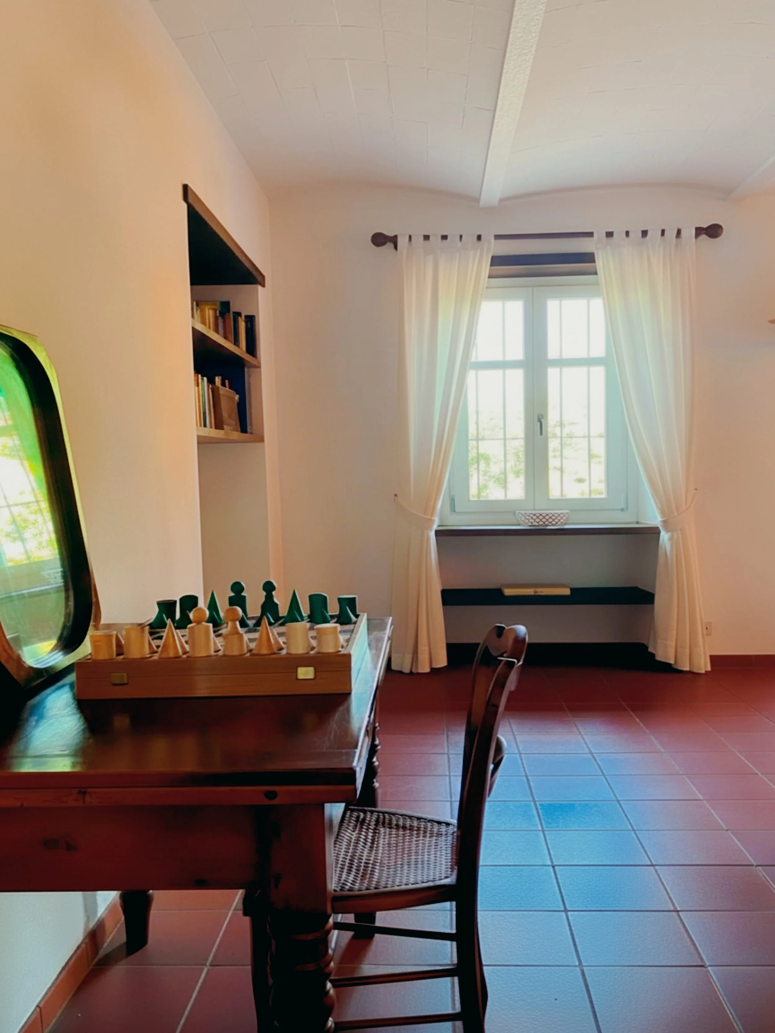 A room with a window covered by white curtains, a wooden table with a chess game, and a bookshelf on the wall.