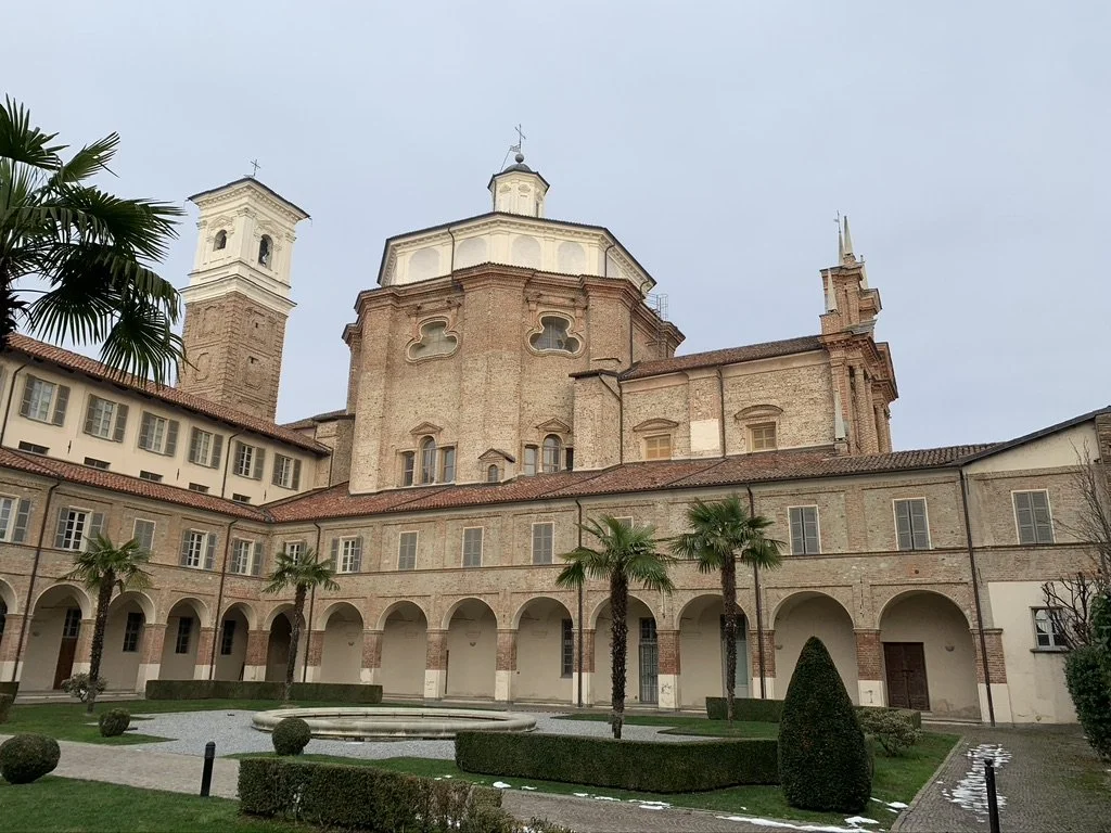A historic church with a large central dome, twin bell towers, surrounded by a courtyard with palm trees and manicured bushes.