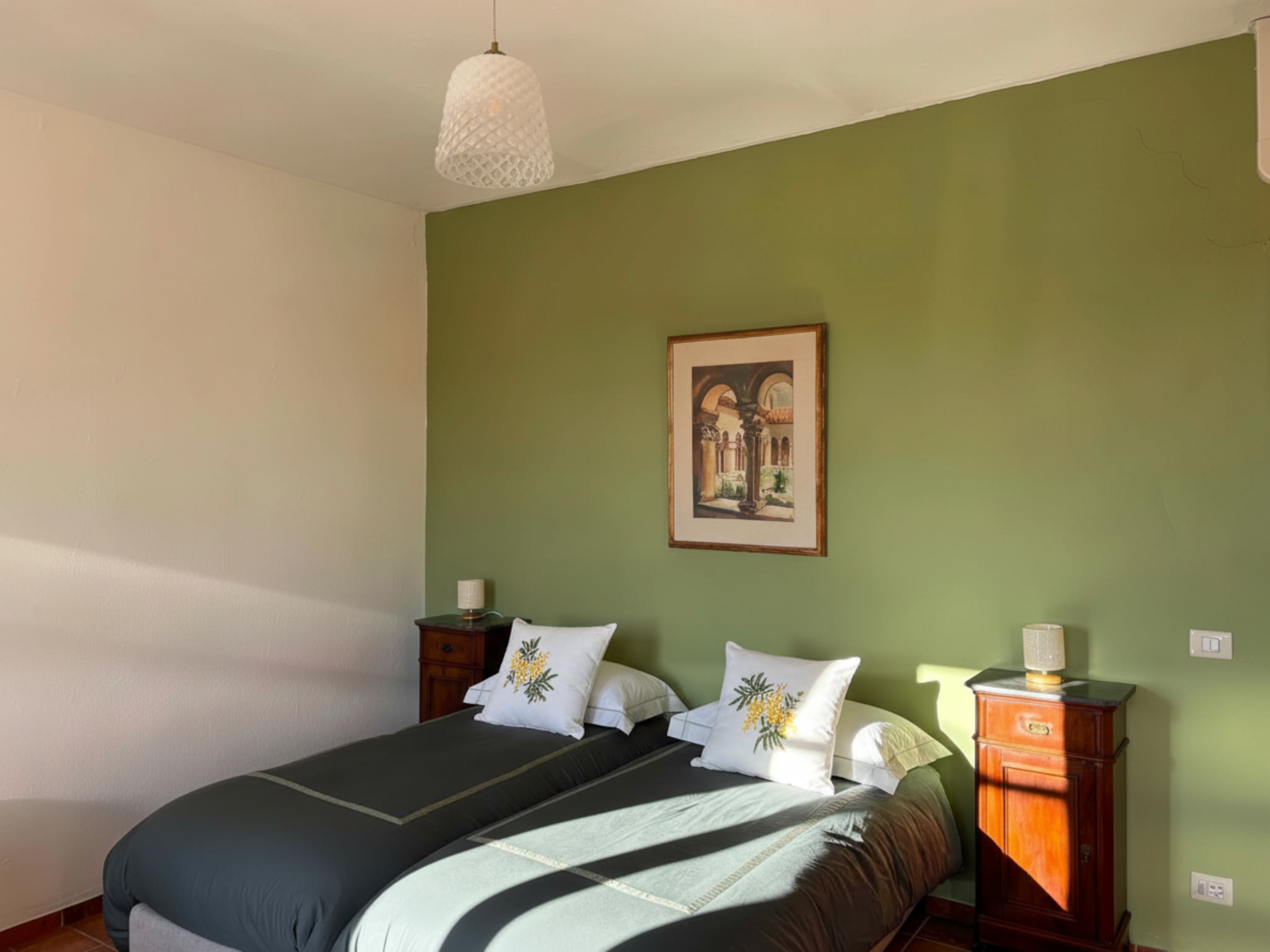 A bedroom with a bed covered in a black and green blanket, decorative pillows with botanical prints, a wooden nightstand with a lamp, a green accent wall, a framed painting, and a white ceiling light fixture.