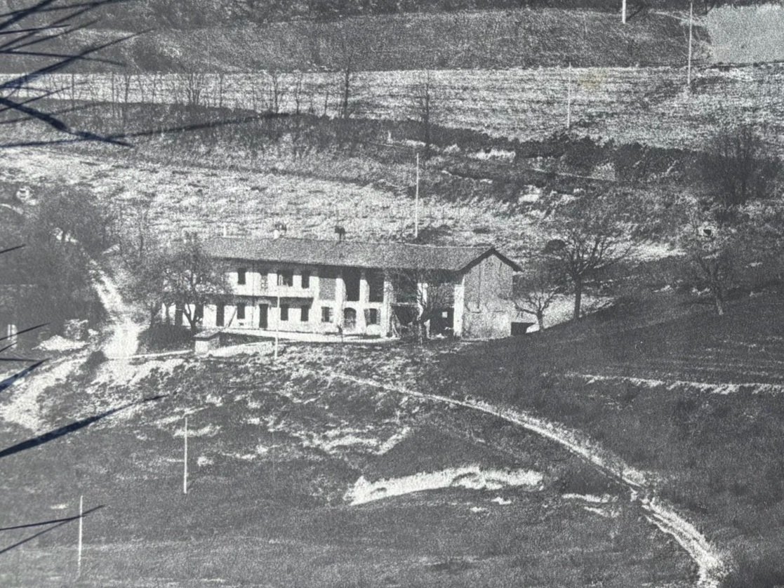 Black and white photo of a rural house with a sloped roof, surrounded by trees and fields, with dirt roads and sparse vegetation in the foreground.