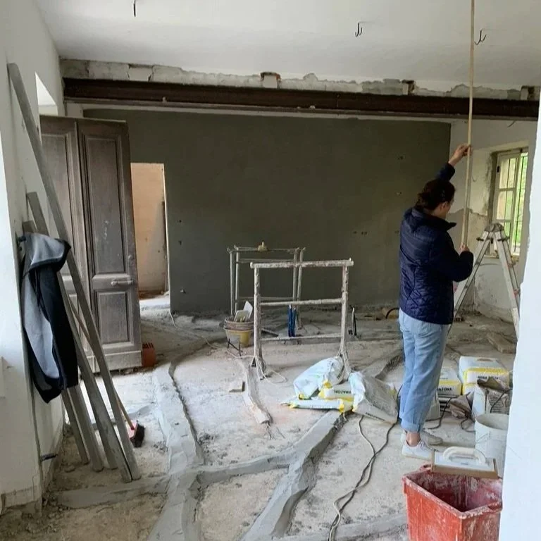 Woman working on renovation inside a room with construction tools and supplies, partially finished walls, and construction debris on the floor.