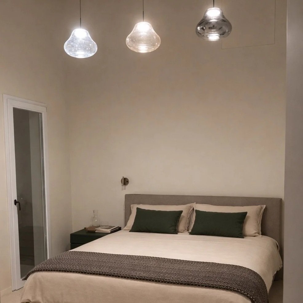 A bedroom with a bed, three pendant ceiling lights, a nightstand with a glass, books, and a wall-mounted reading light.