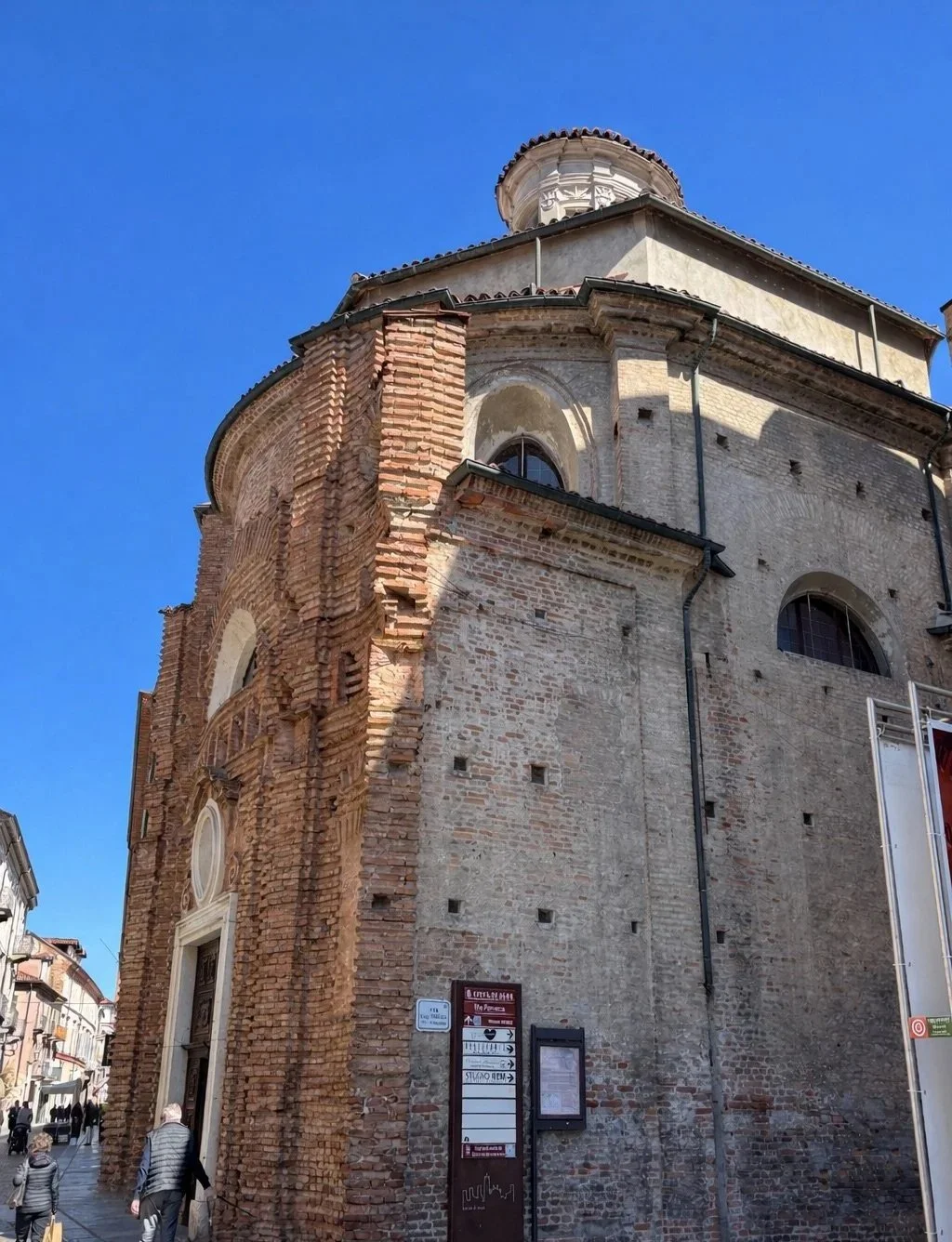 A historic brick and stone church with a rounded top and a small dome, located on a street with people walking nearby, under a clear blue sky.
