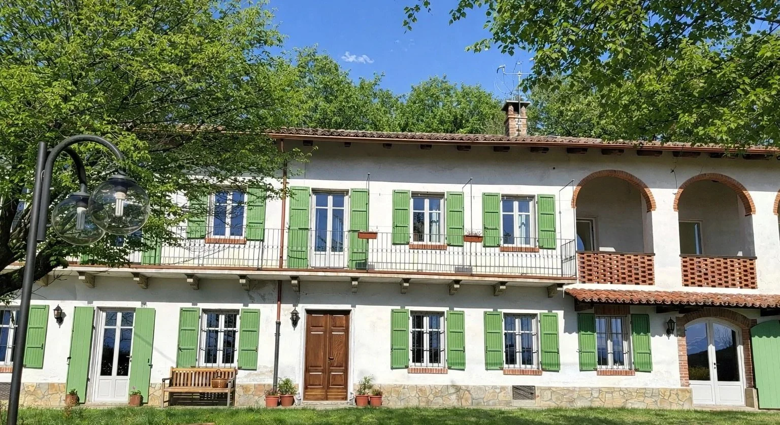A two-story house with white walls, green shutters on the windows, a wooden front door, and a small balcony with brick arches. Green trees surround the house, and a lawn with a bench, potted plants, and a street lamp are in front.