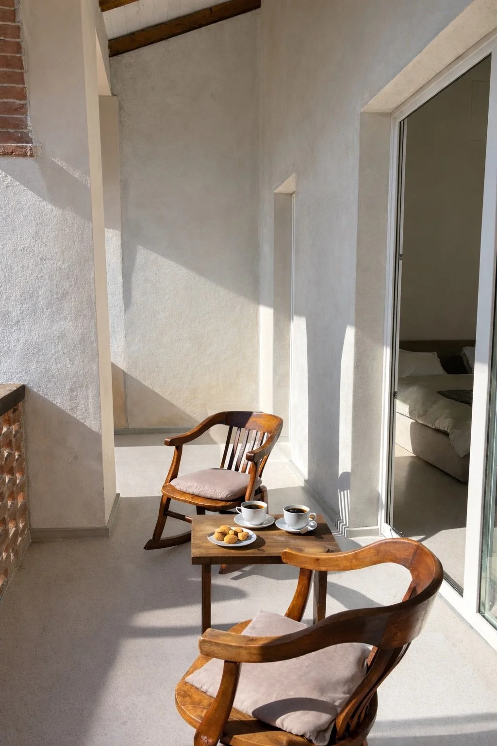 A cozy balcony with two wooden chairs, a small wooden table, two cups of coffee, and a plate of cookies, overlooking a bedroom through a sliding glass door, with sunlight and shadows creating a warm ambiance.