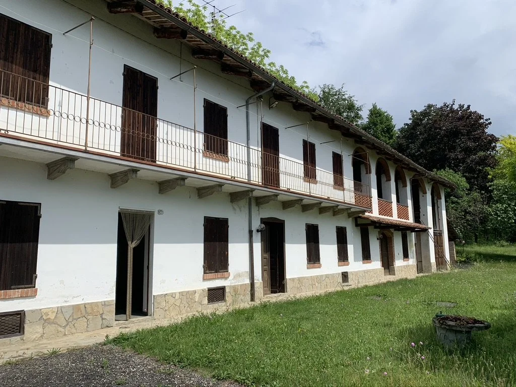 A two-story white building with brown window shutters, a stone foundation, and a balcony on the upper floor. The building is surrounded by green grass and trees, with an overcast sky in the background.