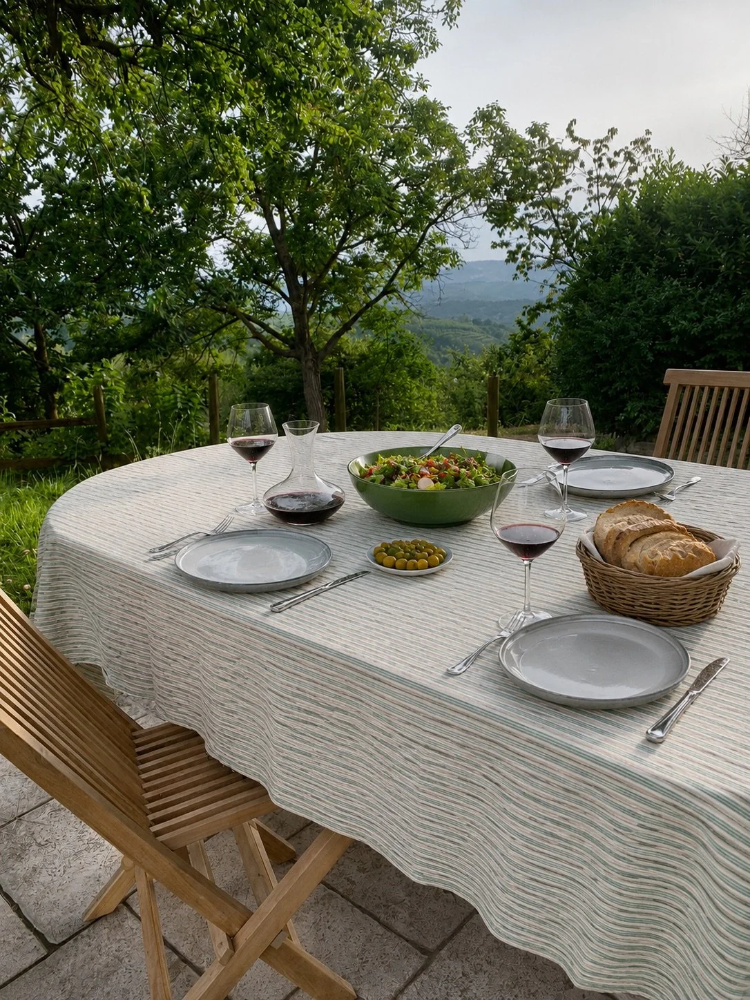 Outdoor dining table with plates, glasses of red wine, a salad, bread in a basket, an olive dish, and utensils on a striped tablecloth, set against a scenic view of trees and mountains.