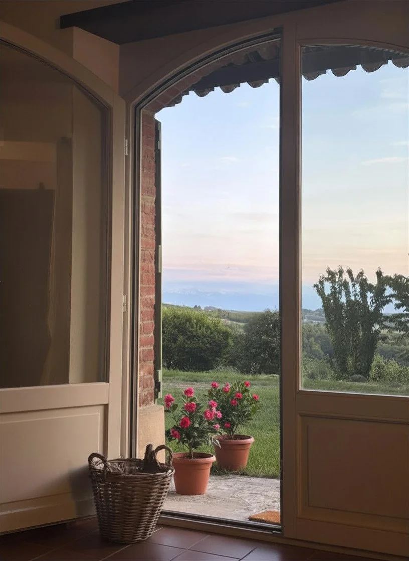 Open door leading to a backyard with pink flowers in pots, green grass, and rolling hills in the distance during dusk or early evening.