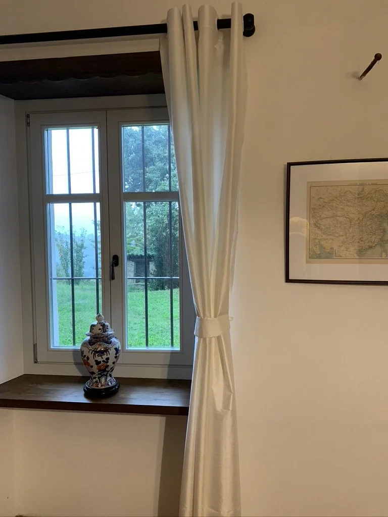 A window with metal bars in a white frame, pulled back cream-colored curtains on a rod, showing a green outdoor scene with trees. A decorative ceramic vase with a floral pattern is on the windowsill, and a framed map hangs on the wall to the right.