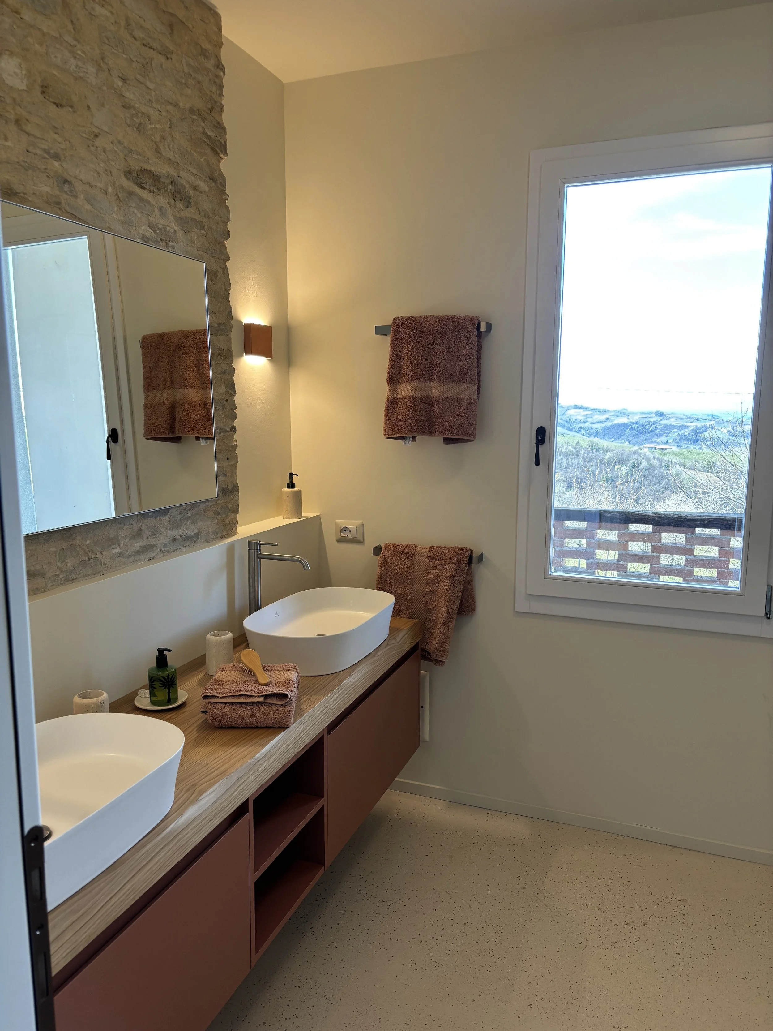 Modern bathroom with a double sink vanity, large mirror, stone wall accent, pinkish-brown towels, soap dispenser, and a large window showing a scenic view of mountains and sky.