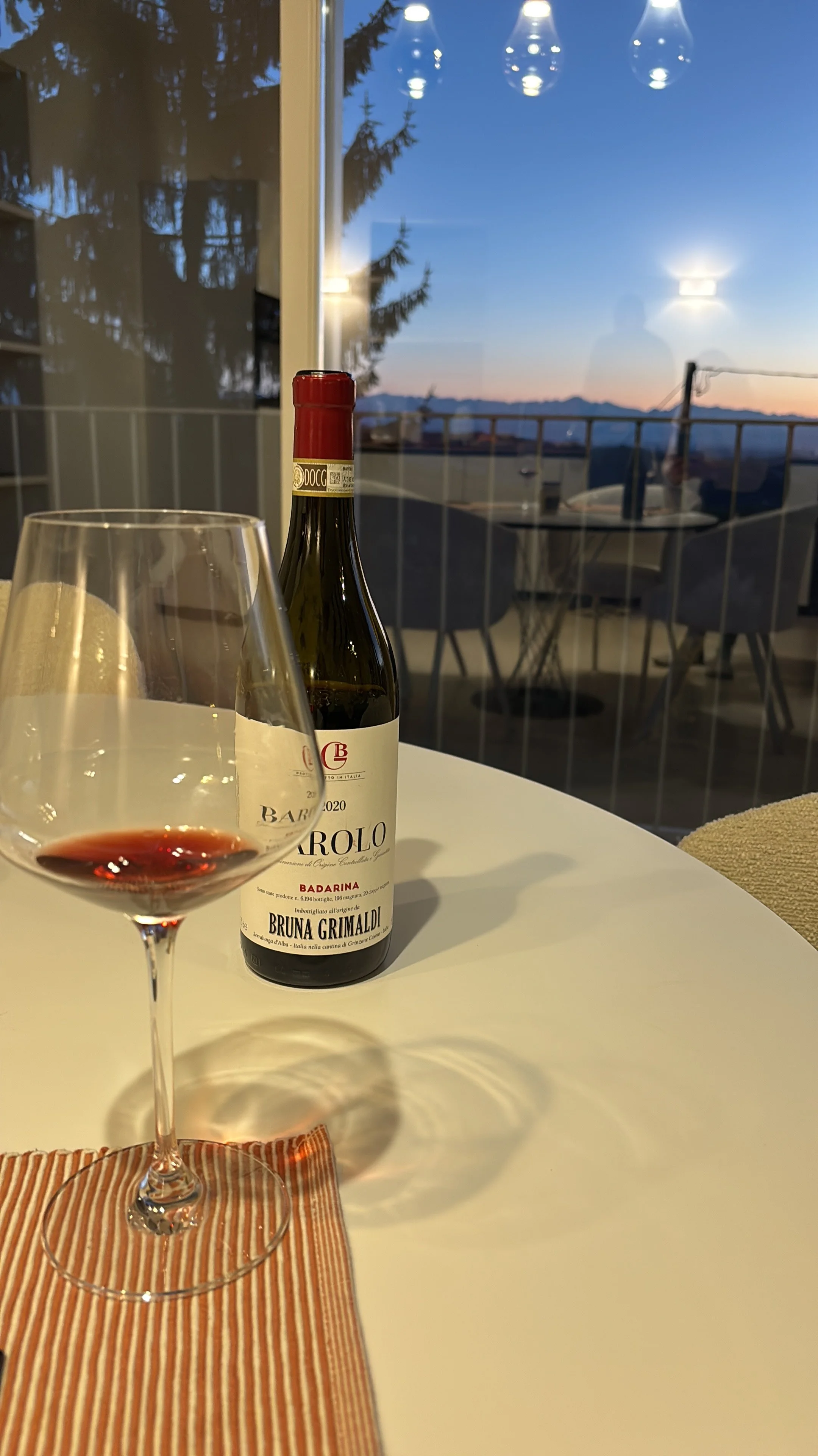 A glass of red wine on a table with a striped cloth, a bottle of red wine, and a blurred background of a balcony and a scenic sunset view.