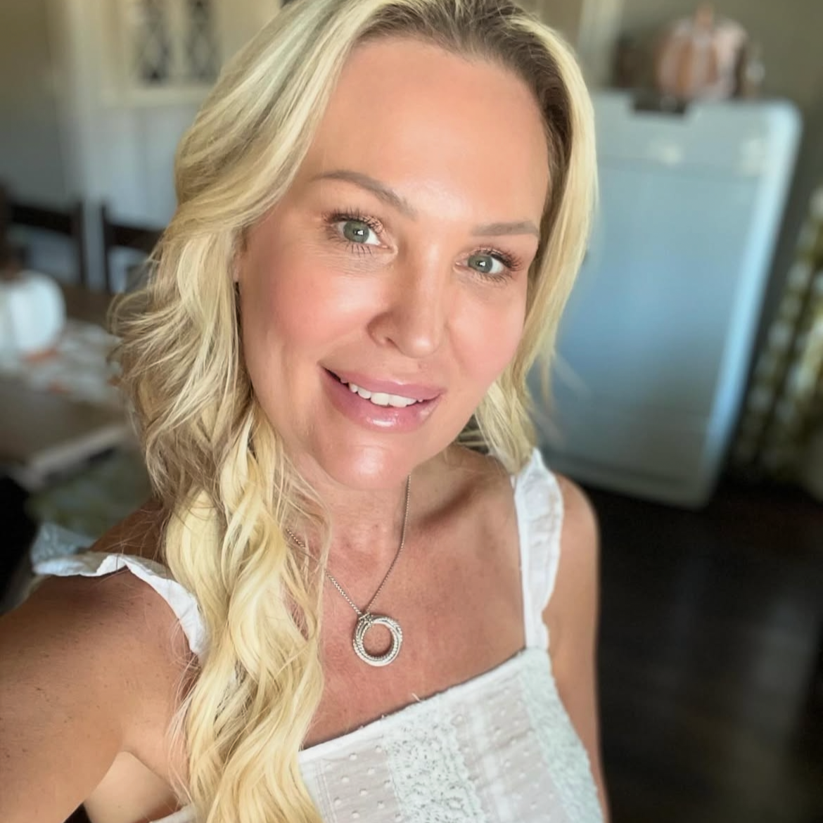 A woman with long blonde wavy hair and green eyes smiling while taking a selfie indoors. She is wearing a white top with ruffled shoulder straps and a circular silver necklace.