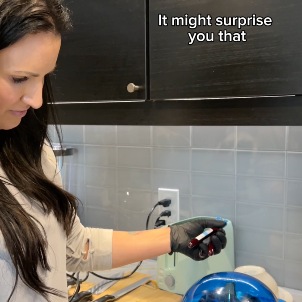 A woman with long dark hair, wearing a beige shirt and black gloves, is in a kitchen, holding a test tube with a red liquid. There are kitchen cabinets and a white electrical outlet in the background.