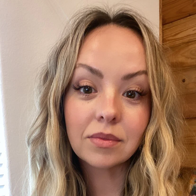 Close-up of a young woman with long wavy blonde hair, light makeup, and a neutral expression, standing indoors near a wooden wall.