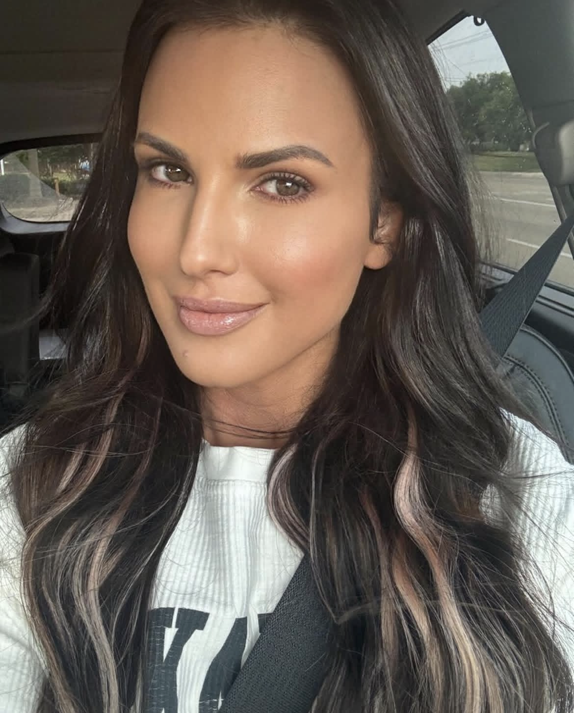 A woman with long, wavy dark hair and light makeup sitting in a car, wearing a white top and a seatbelt.