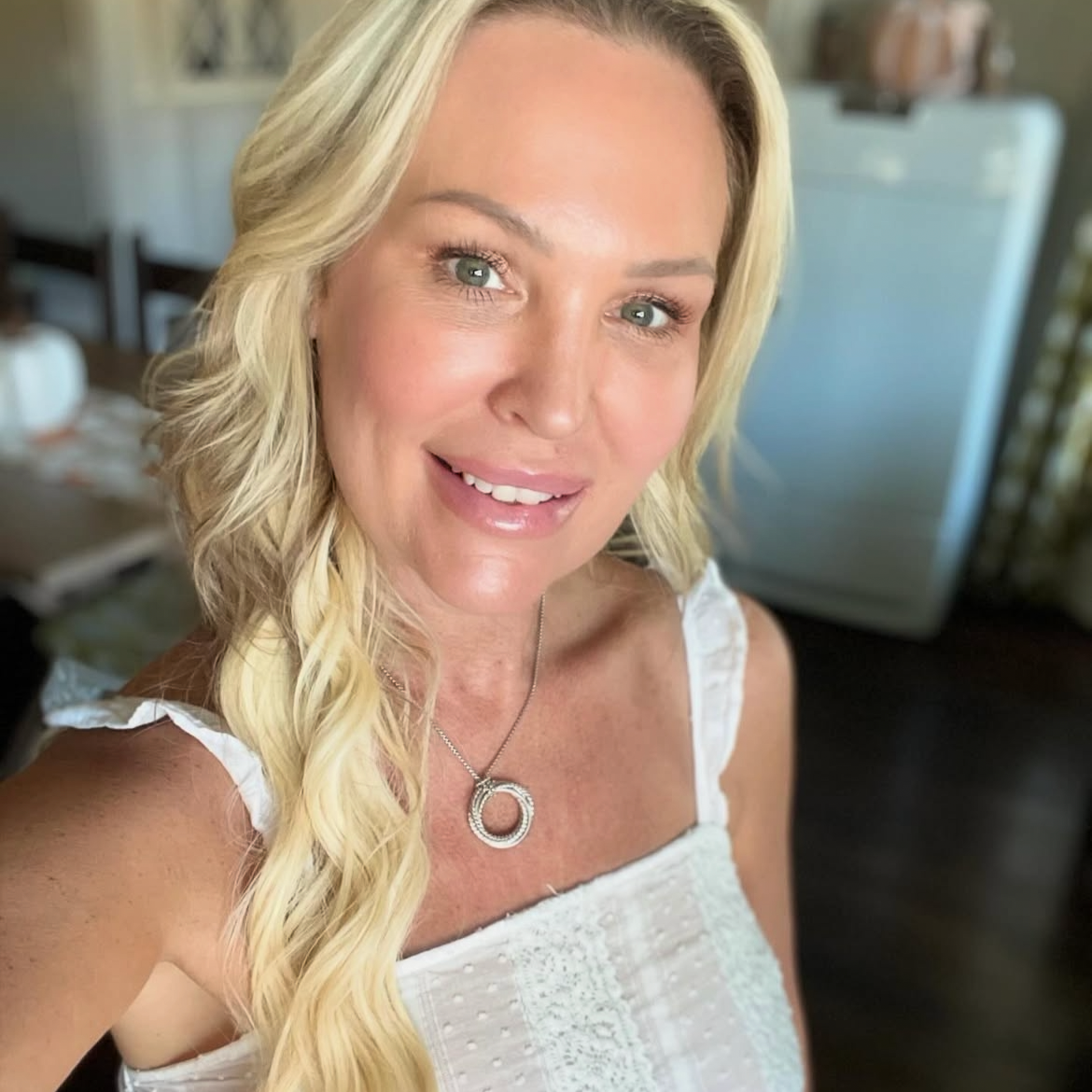 A woman with long blonde hair, wearing a white top with ruffled straps and a circular necklace, smiling at the camera.