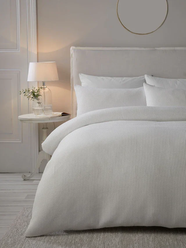 A neatly made bed with white bedding and pillows in a minimalist bedroom with a bedside table, lamp, and a small decorative plant.