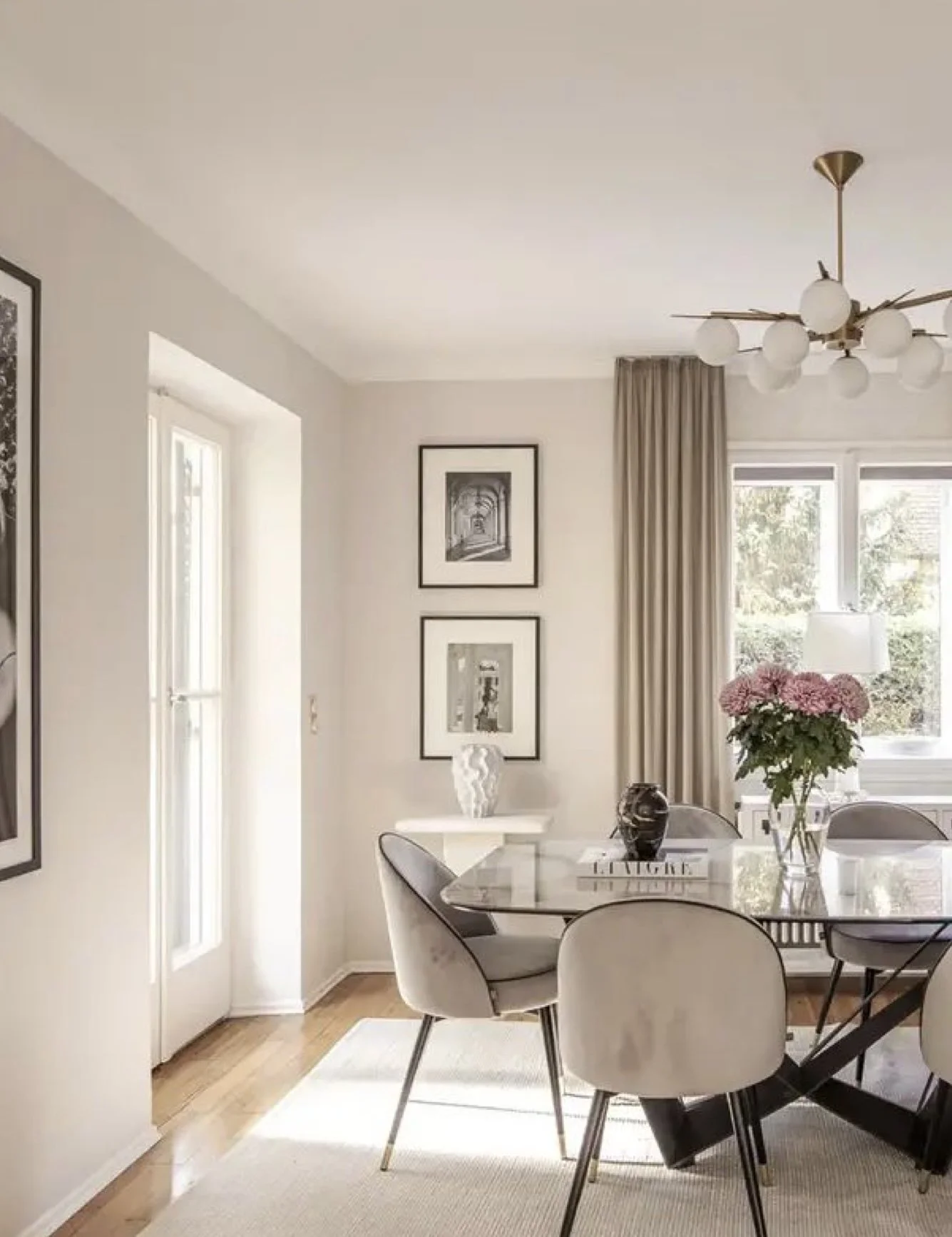 A dining room with a glass table, surrounded by cream-colored chairs, decorated with pink flowers in a vase, and artwork on the walls. The room has a window with beige curtains and a modern ceiling light fixture.