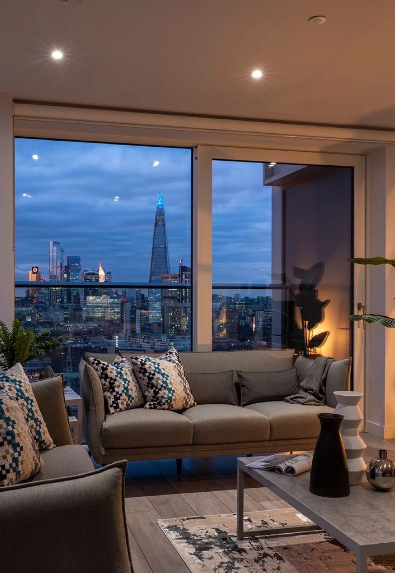 Living room with gray sofa and patterned throw pillows, a small white coffee table with decorative vases, and a large window showing a city skyline at dusk with tall buildings, including The Shard in London, illuminated against a cloudy sky.