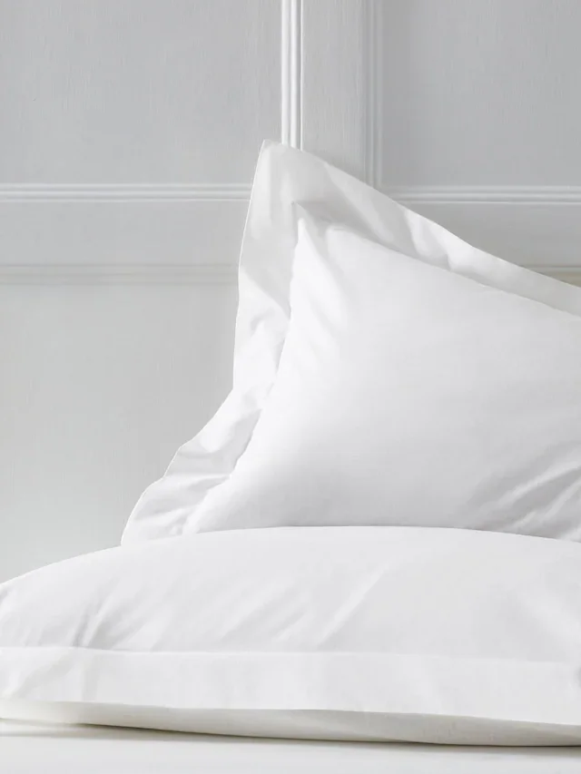 Close-up of a white pillow on a bed with white bedding and a white paneled wall in the background.