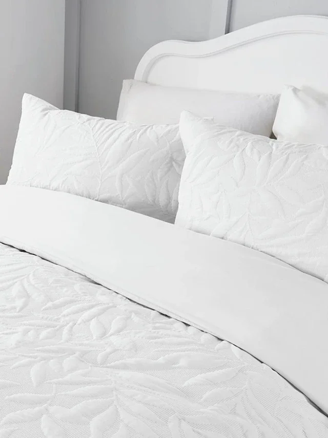 A neatly made bed with white bedding, including pillows and a quilted bedspread, in a minimalist bedroom.