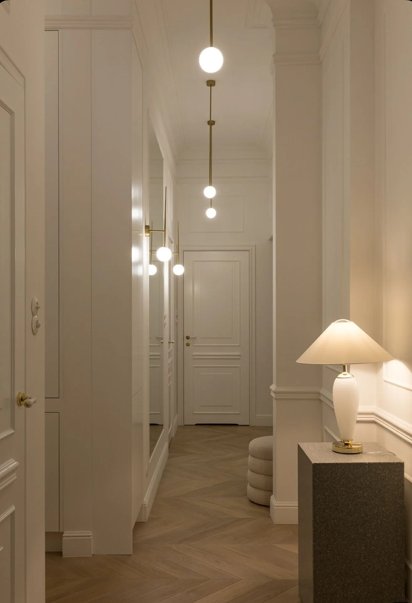 Elegant hallway with white walls, wooden herringbone flooring, and modern lighting fixtures including a ceiling bulb string and a table lamp on a granite pedestal.