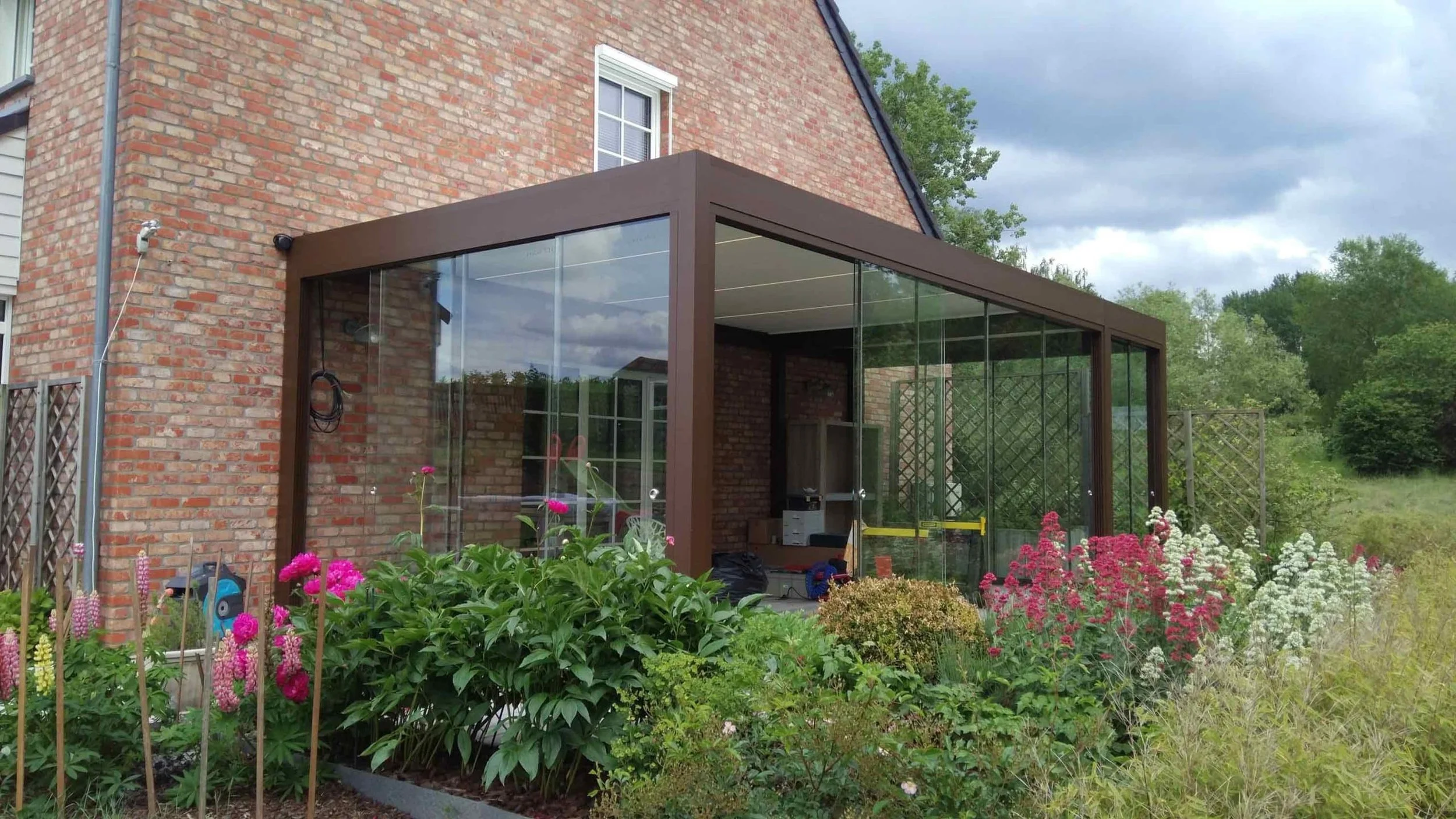Een tuin met kleurrijke bloemen en een afgesloten veranda met glazen wanden achter een rood bakstenen huis, onder een bewolkte hemel.