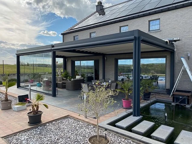 Een overdekte patio met een zitgedeelte onder een modern glazen dak, terras met potted planten en een klein vijvertje met leisteen stappen.