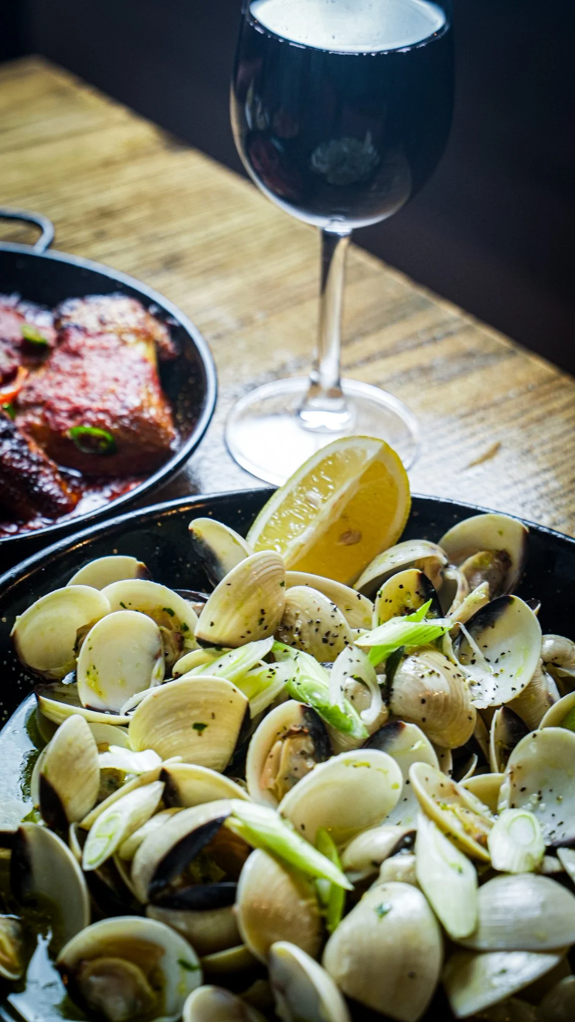 A bowl of cooked clams garnished with chopped green onions and black pepper, with lemon wedges on the side, a glass of red wine, and a dish of cooked meat with sauce on a wooden table.
