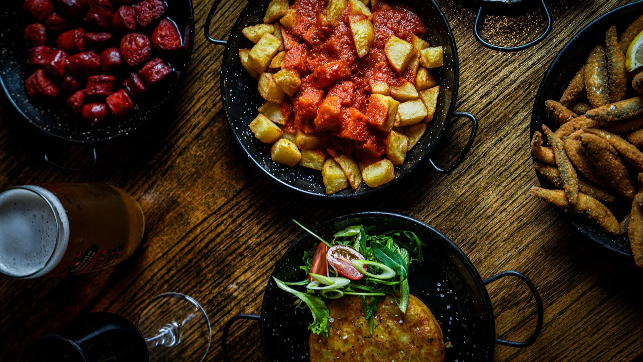 A top-down view of a wooden table with various dishes including a bowl of cherry tomatoes, a plate of roasted potatoes with tomato sauce, a salad with greens and cherry tomatoes, fried chicken wings, and beverages such as beer and a dark soda.