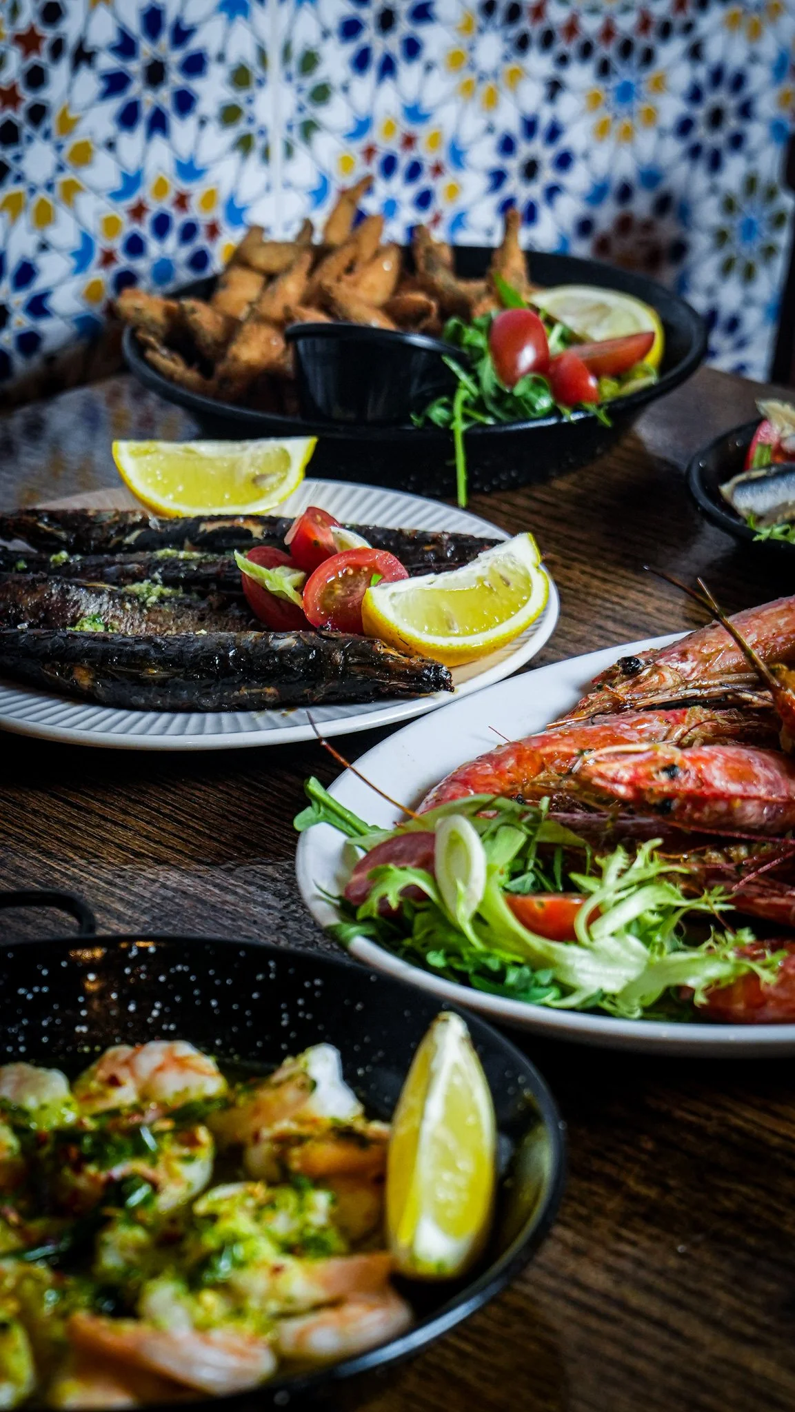 A variety of seafood dishes on a wooden table, including grilled fish with lemon wedges, prawns, shrimp salad, and fried chicken wings with salad.