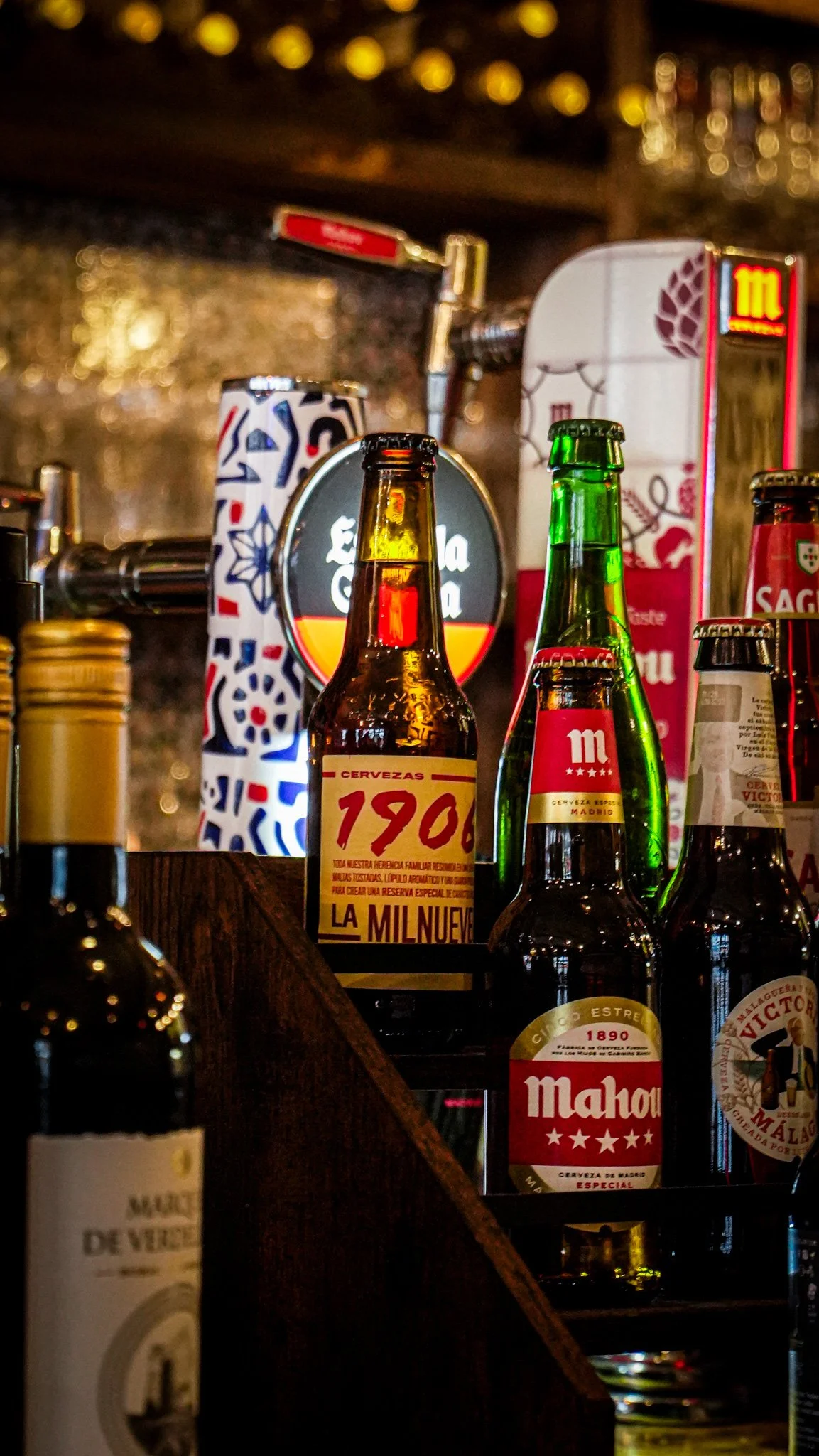 Several glass bottles of beer displayed on a bar shelf, with labels including La Mil Nuev, Mahou, and others, illuminated with warm lighting.