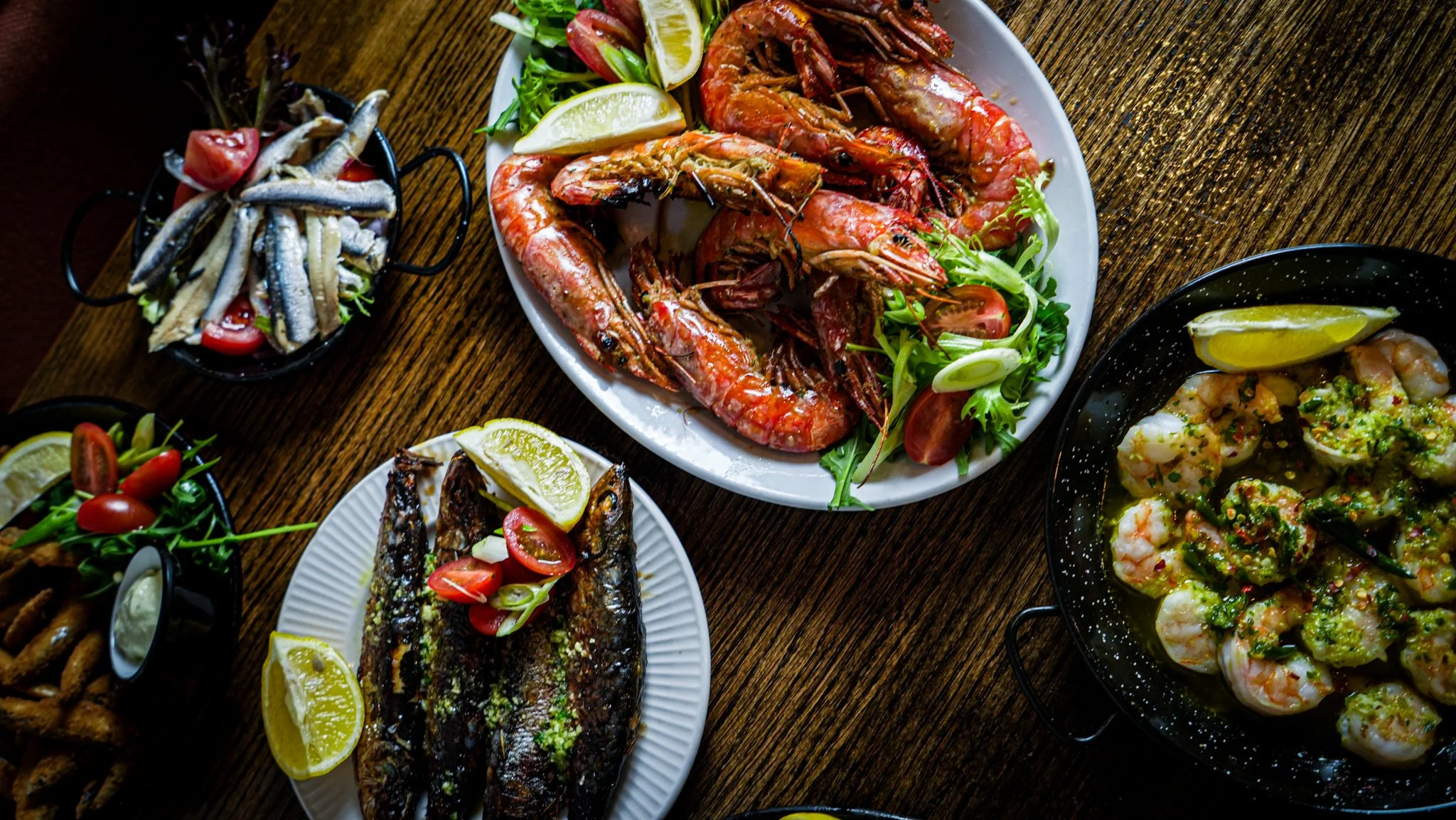Various seafood dishes on a wooden table, including a plate of grilled shrimp with lemon wedges and salad, a whole roasted fish garnished with lemon and cherry tomatoes, a black skillet of peeled shrimp in sauce with lemon, and a small bowl of small 