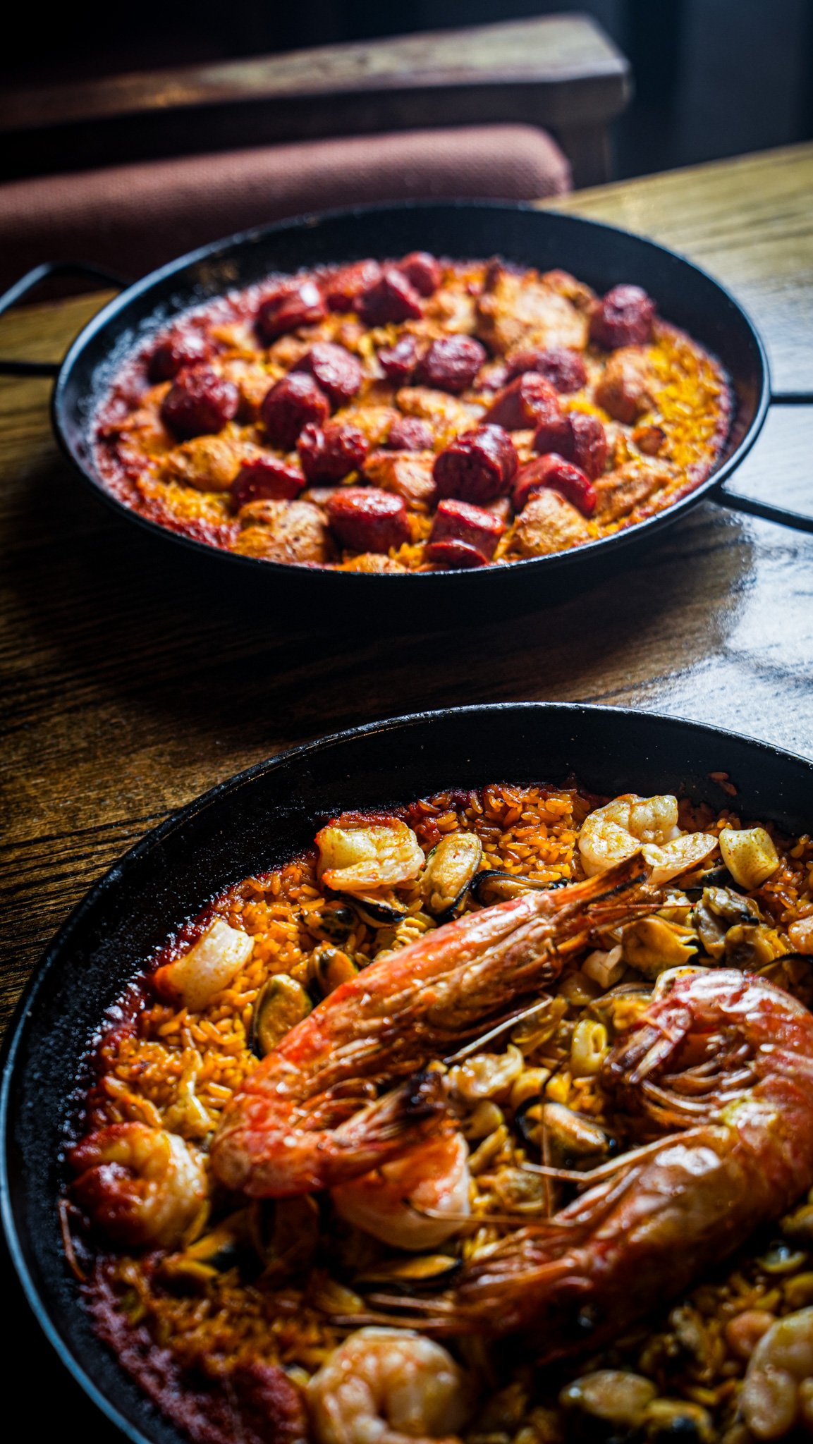 Two black skillets on a wooden table filled with seafood paella. The front skillet has shrimp, clams, and rice, while the back skillet contains chicken and chorizo with rice.