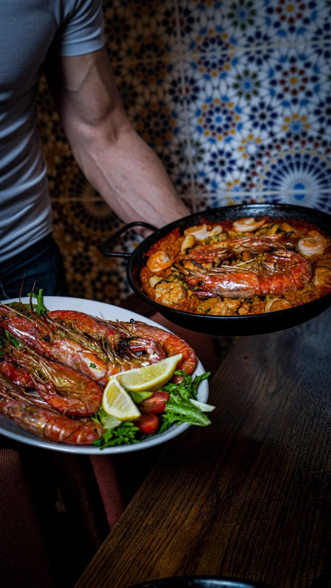 A person holding a black pan filled with shrimp and seafood in a red sauce, with a decorative blue and white patterned wall in the background.