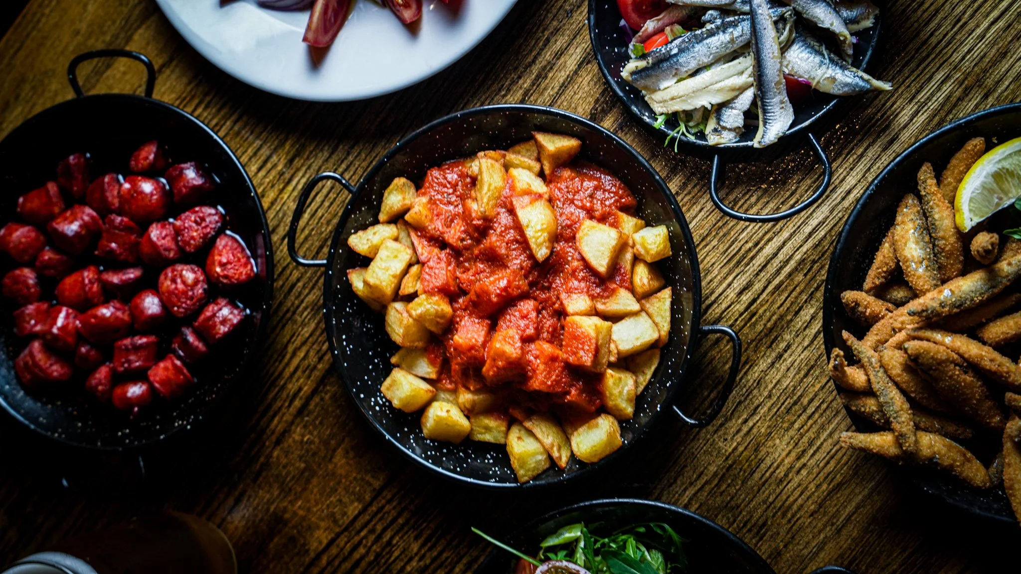 Assorted dishes on a wooden table: a bowl of cooked potatoes with tomato sauce, a bowl of fried chicken strips with lemon, a plate of cherry tomatoes, a plate with mixed fish fillets and vegetables, and a plate of fried small fish.