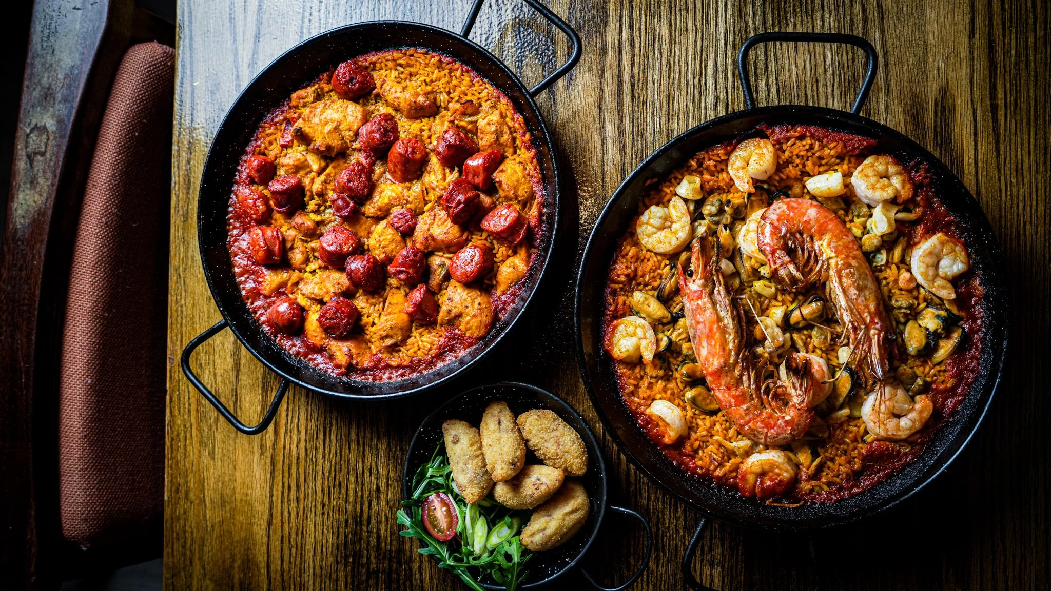 Two pans of seafood and chicken paella with rice, shrimp, and mussels, accompanied by fried chicken tenders, cherry tomatoes, and greens on a wooden table.