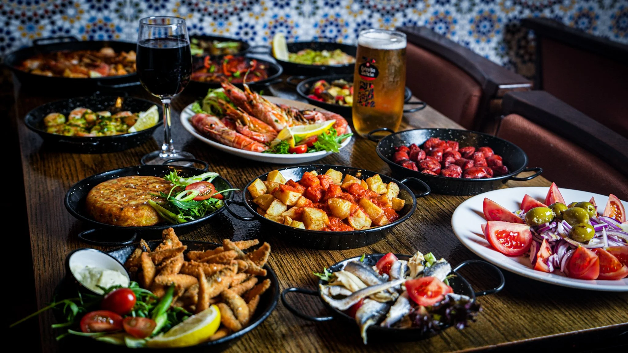 A table filled with various dishes including grilled shrimp, a tomato and lettuce salad, fried fish, roasted potatoes, meatballs, and other Mediterranean foods, with drinks including a glass of red wine and a beer.