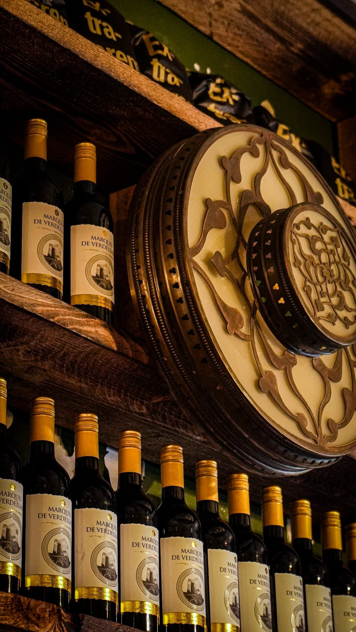 Shelf with bottles of Marqués de Vadelia wine and decorative wall clock in a wooden setting.