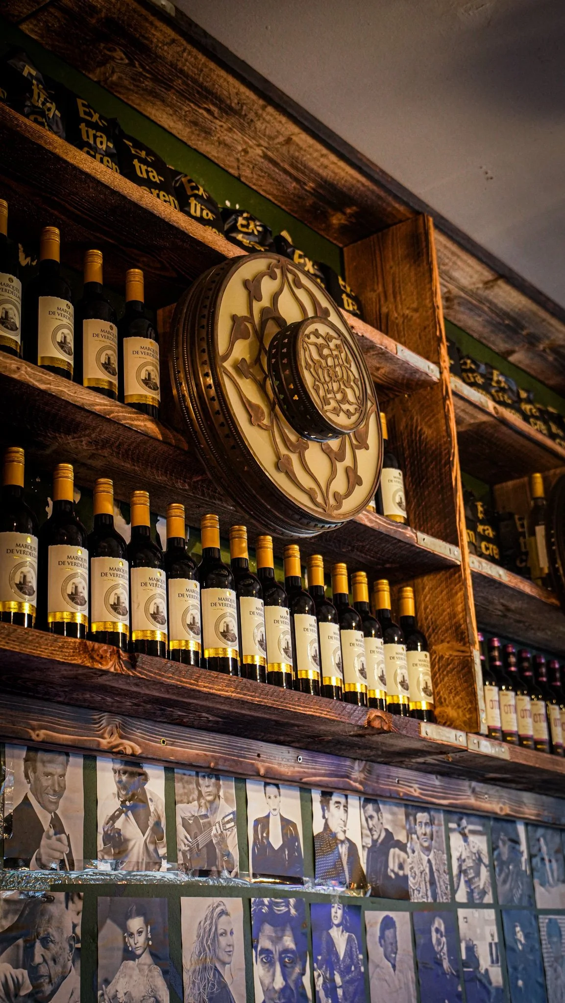Shelf with multiple bottles of wine, a decorative vintage clock, and black ribbons with yellow text, with black and white photographs of various celebrities on the wall.