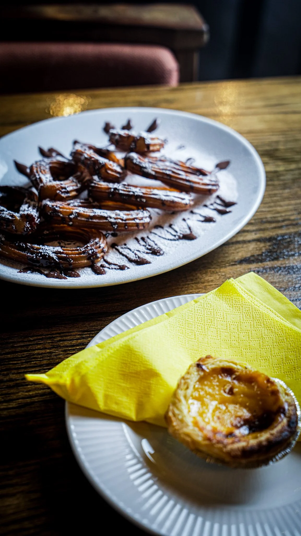 Chocolate-covered waffles with powdered sugar on a white plate, and a partially eaten egg tart on a small white dish with a yellow napkin, on a wooden table.