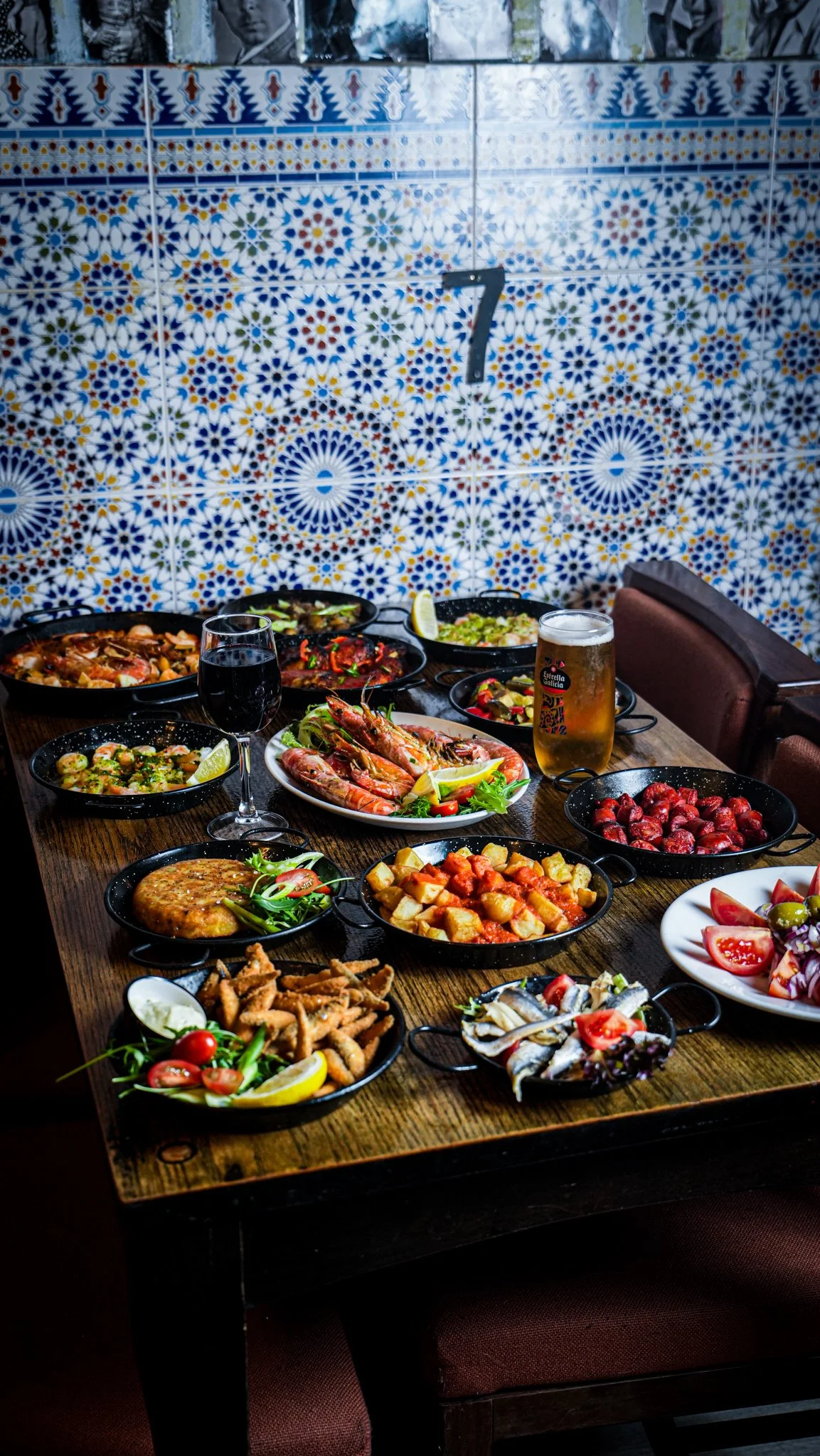 Various Spanish dishes on a wooden table in a restaurant with colorful tile wall in the background.