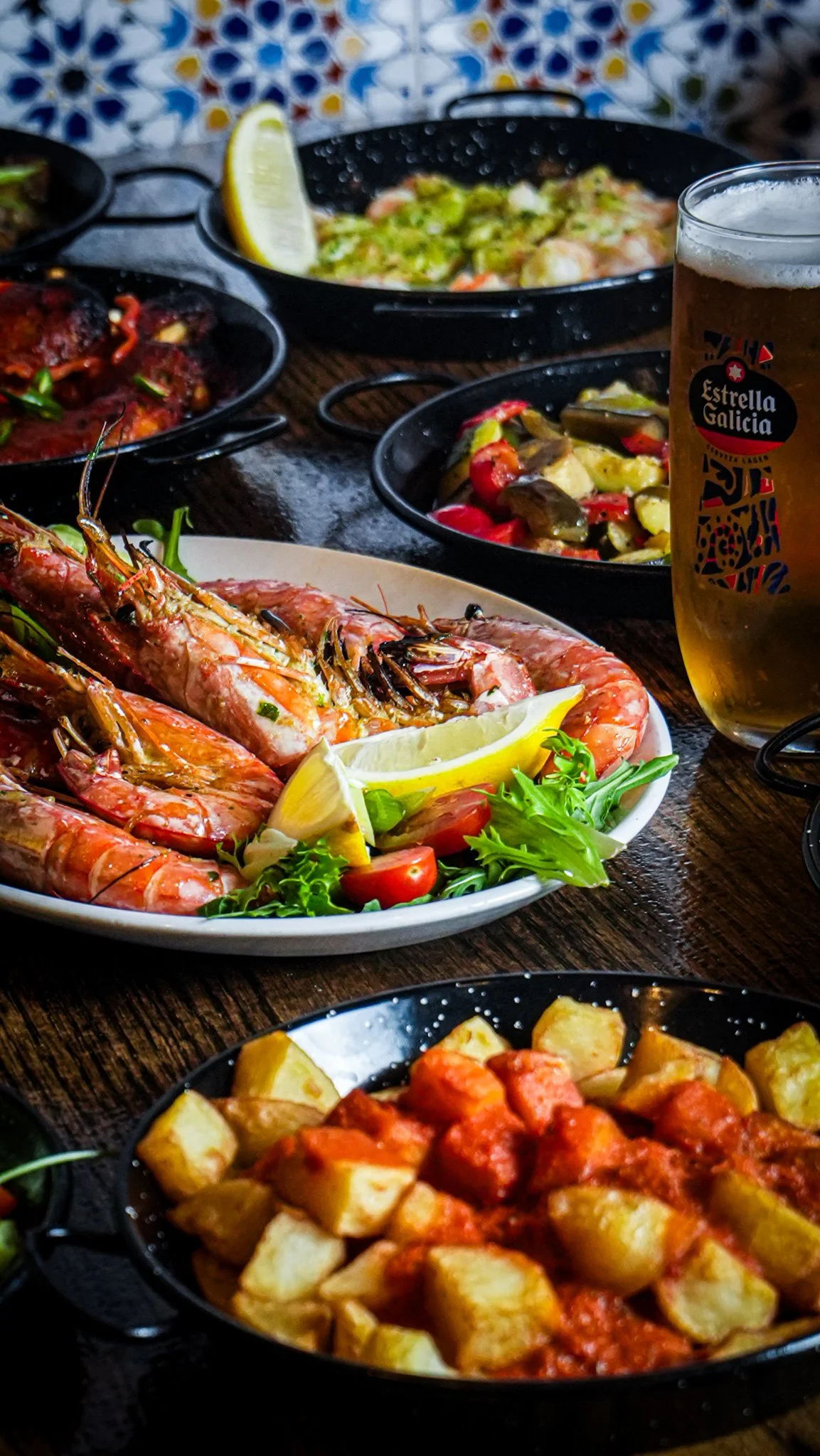 Seafood dishes including grilled prawns with lemon wedges, vegetable salads, and a glass of beer on a wooden table.