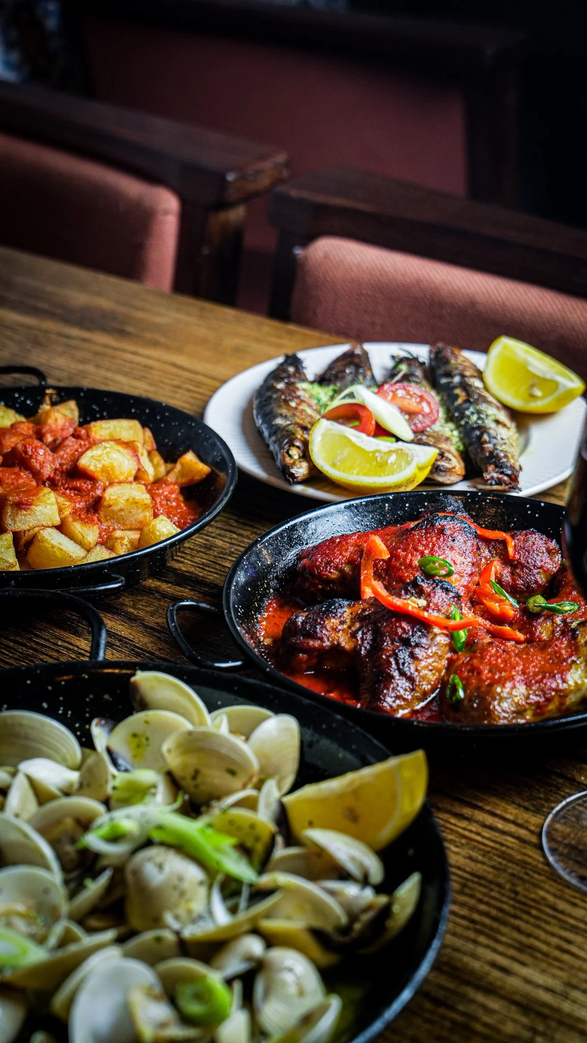 A table with various dishes of seafood and sides, including grilled fish with lemon, clams, potatoes, and a meat dish with sauce.