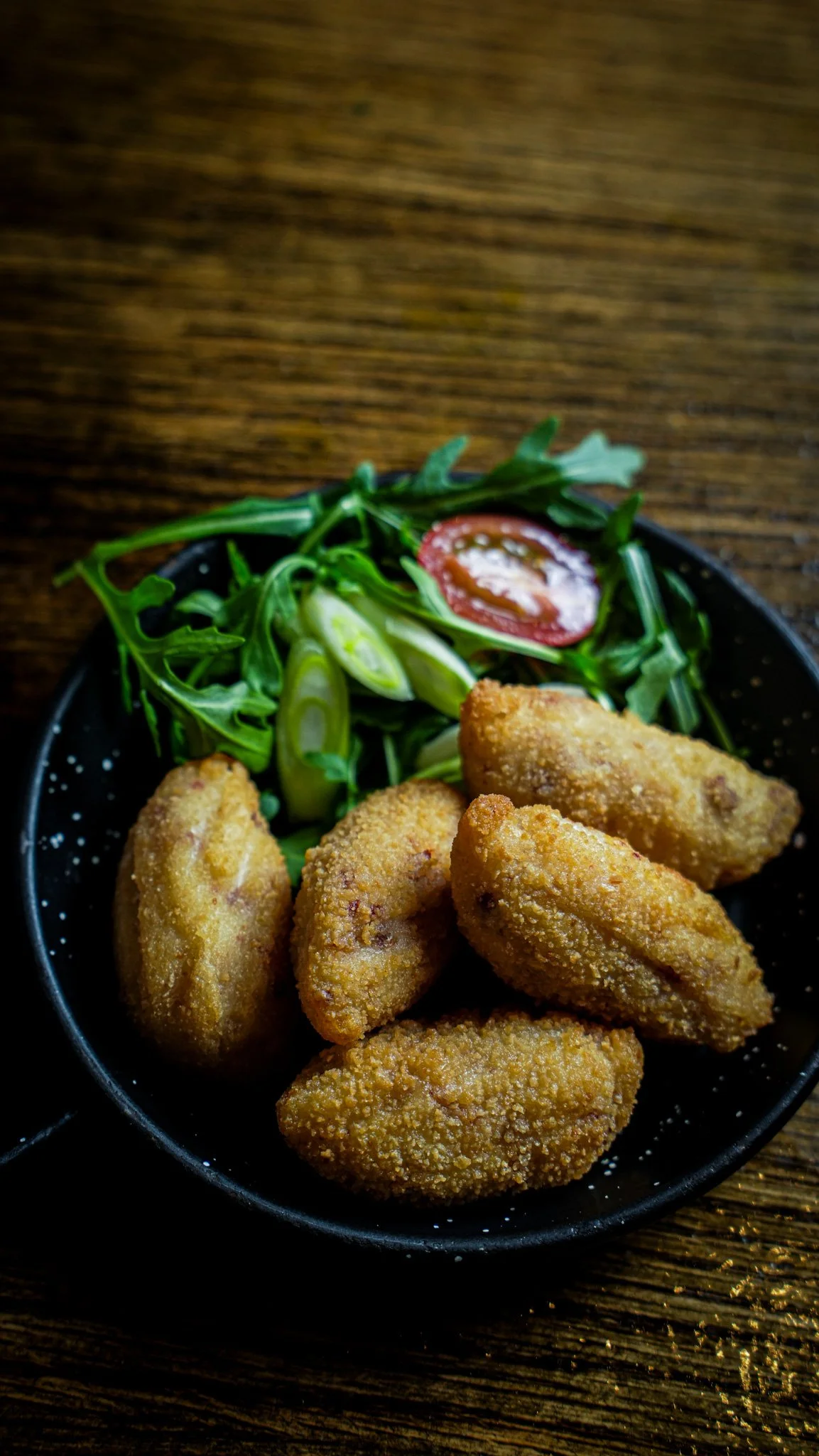 Bowl of fried chicken nuggets with a side salad of arugula, sliced green onions, and a cherry tomato, served on a wooden table.