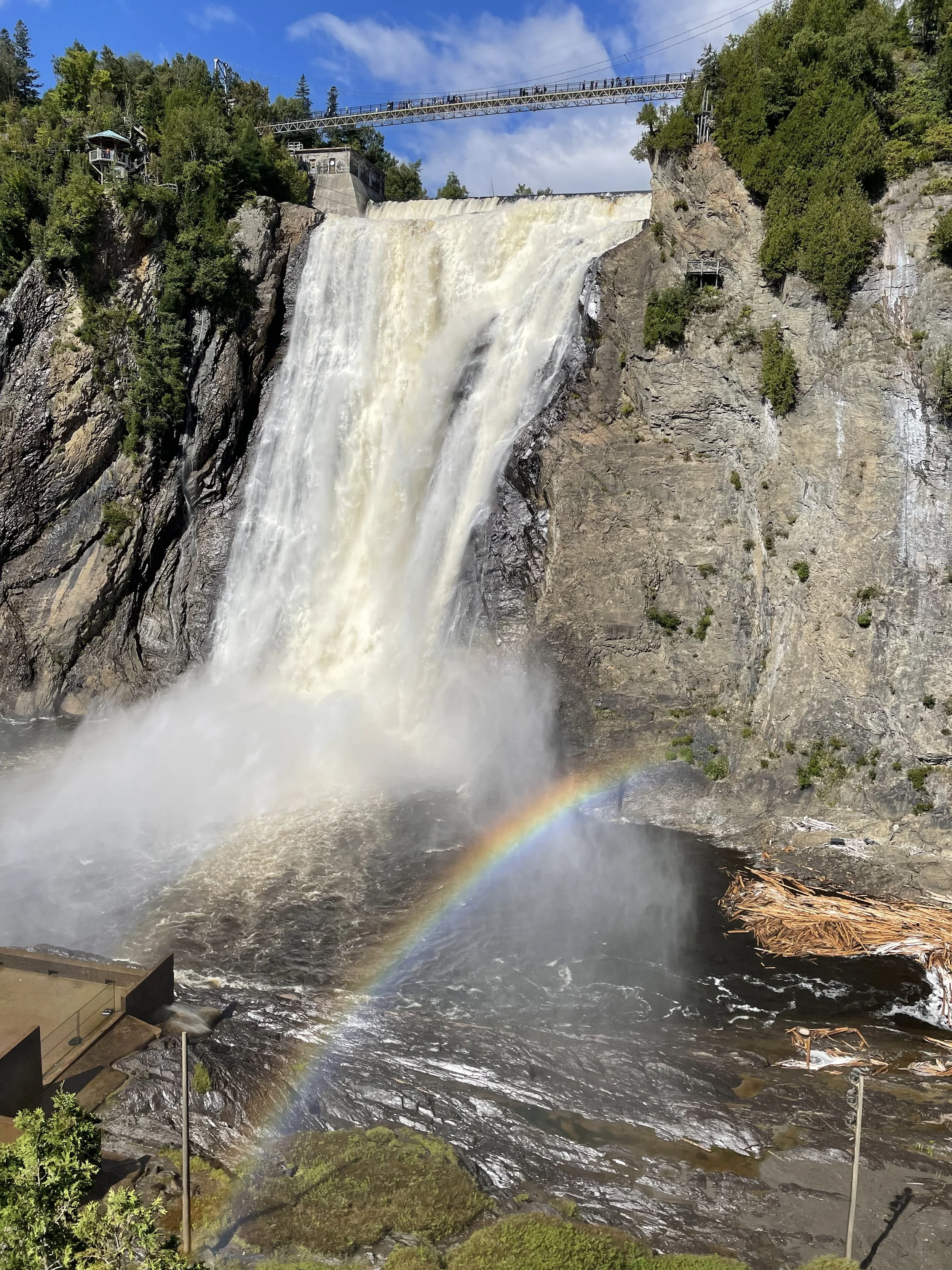 A large waterfall cascading down rocks with a rainbow at the bottom and a small rainbow mist in the foreground. A bridge with people crossing is above the waterfall, surrounded by trees and blue sky with clouds.